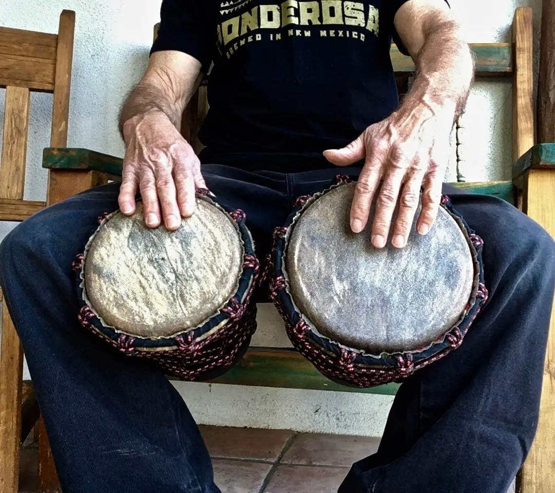 A man sitting on a wooden bench with two drums, playing the Ilimba.