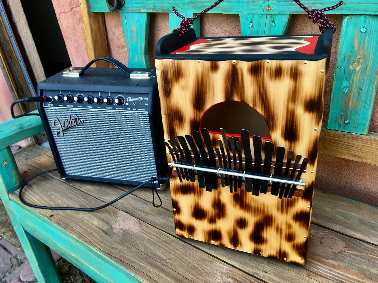 A wooden box with a guitar and amp, accompanied by an African drum, sitting on a wooden bench.