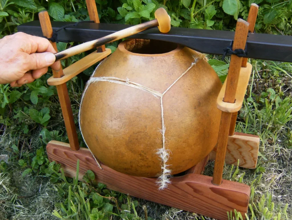 A person is holding a wooden pot, resembling a gourd instrument, on a wooden stand.