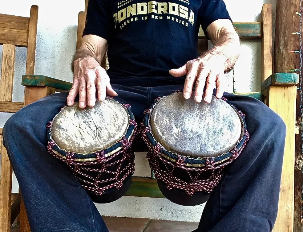 A man sitting on a wooden bench with two djembes, playing African harps.