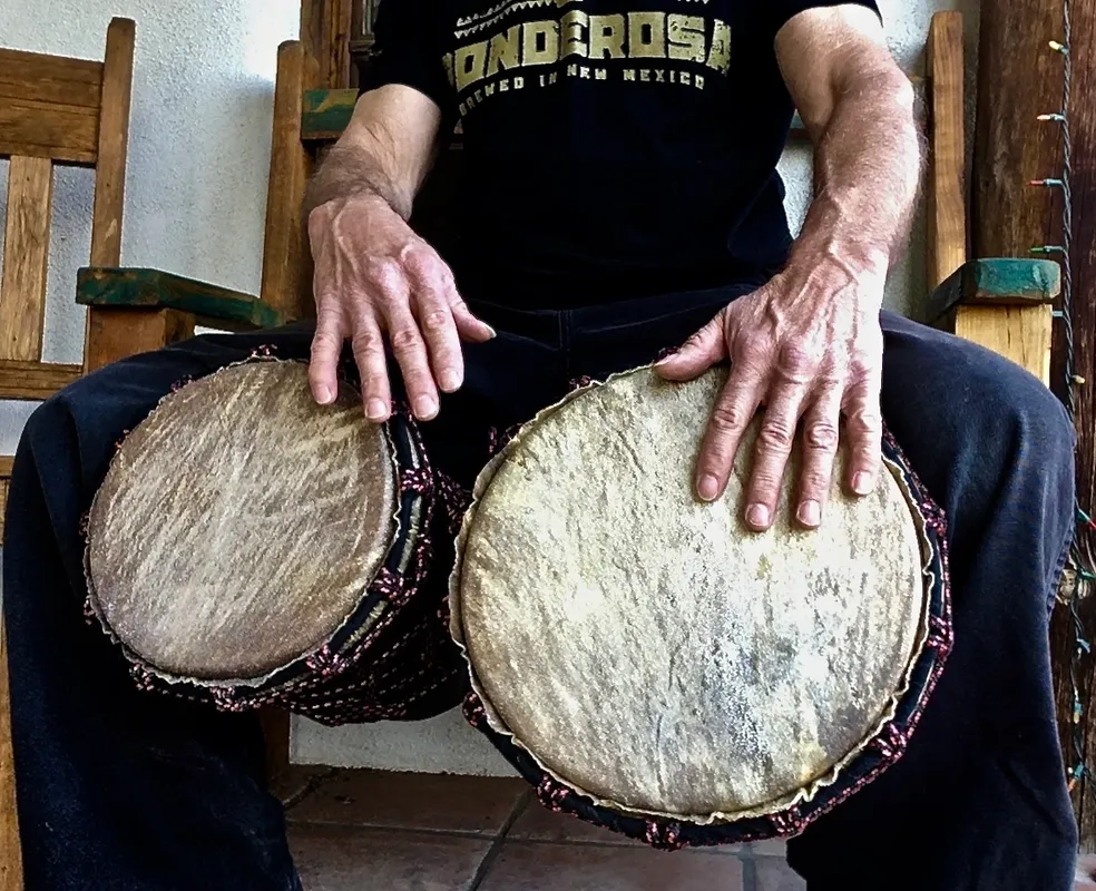 A man sitting on a wooden bench with two drums, playing the African harps.