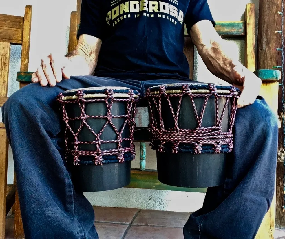 A man sitting on a wooden bench with two drums, playing African harps.