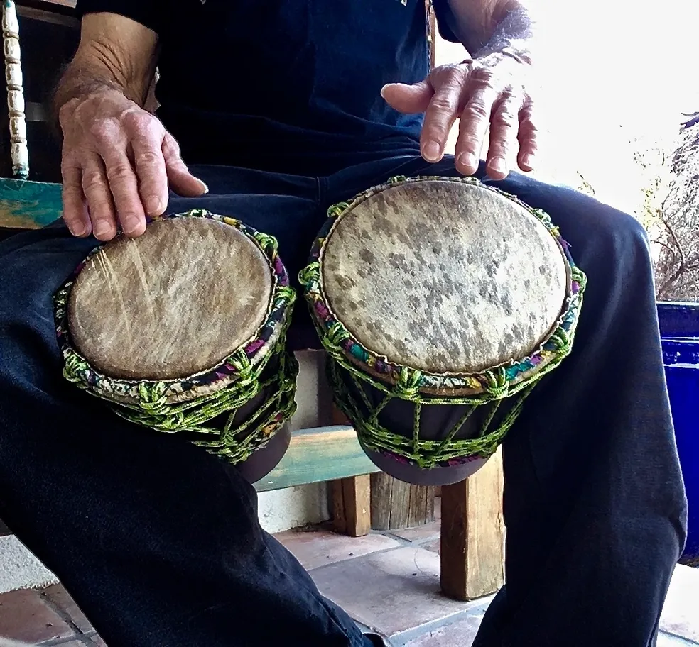 A man sitting on a bench with two djembes, playing the African harps.